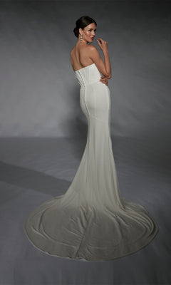 A woman wears the Long White Dress: Alyce 7102, featuring a minimalist design with a strapless corset bodice and long train, as she stands against a gray backdrop, facing away and looking over her shoulder.