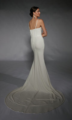 A woman stands facing away against a dark background, wearing the Long White Dress: Alyce 7104—a fitted formal gown with a long train.