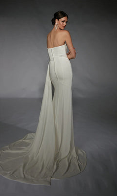 A woman models the Alyce 7105, a strapless long white dress with a train and button details, standing against a gray backdrop and looking over her shoulder.