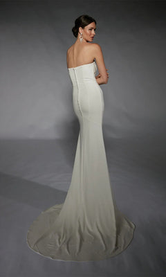 A woman faces away from the camera, wearing the Alyce 7105 long white dress—a strapless formal gown with a fitted bodice and flowing train—against a plain gray background.