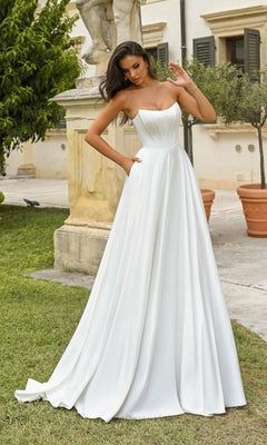A woman stands outdoors near a stone statue and potted plants wearing the Alyce 7110 long dress in Diamond White, featuring a fitted bodice, full satin A-line skirt, and elegant strapless formal design.