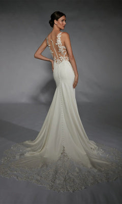 A woman poses in the Alyce 7117 long white dress, featuring a fitted silhouette, ivory hue, sheer back, floral lace accents, fabric-covered buttons, and a long lace train against a gray background.