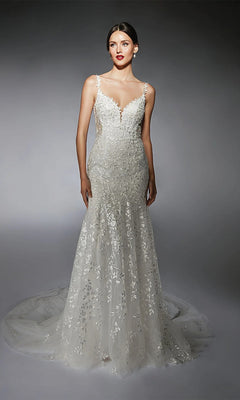 A woman stands against a gray gradient background wearing the fitted, sleeveless Long White Dress: Alyce 7120, featuring a floral lace overlay and a long, trailing train.