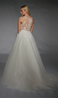 A woman stands facing away against a gray background, wearing the Long White Dress: Alyce 7128, a bridal gown featuring a sheer floral lace appliqué back and a long tulle skirt.