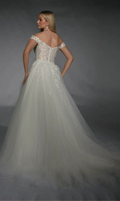 A woman stands facing away against a dark background, wearing the Alyce 7129 long white dress featuring an off-the-shoulder, ivory ball gown design with a lace corset bodice and floral details.