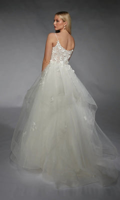 A woman stands against a gray background, facing away from the camera, wearing the Alyce 7131 long white dress—a sleeveless bridal ball gown with a tulle ruffled skirt and 3D floral lace appliques.