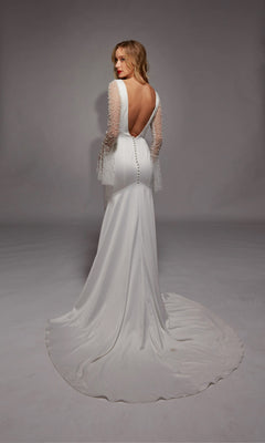 A woman in the Long White Dress: Alyce 7137, featuring illusion lace sleeves and an open back, stands on a plain background with her back to the camera.