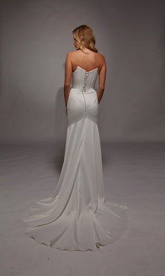 A woman stands facing away against a plain background, wearing the Long White Dress: Alyce 7139—a strapless satin gown with a lace-up back and elegant button details.