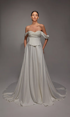 A woman stands in a studio wearing the Alyce 7142 long white dress, featuring an off-the-shoulder satin design with a fitted corset bodice and a floor-length flowing skirt with a train.