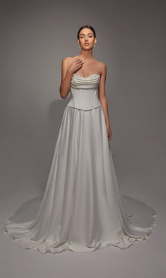 A woman wears the Alyce 7142, a long white dress with a corset bodice and flowing train, its satin fabric softly shimmering against a plain gray background.
