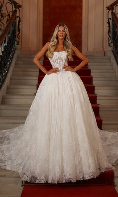 A woman in an ivory solid Long White Dress: Alyce 7149 stands on a grand red-carpeted staircase, hands on her hips, showcasing the elegant ball gown style.