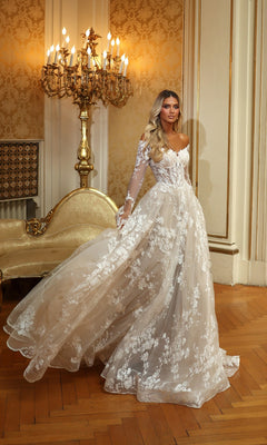 A woman wears the Alyce 7150 long ivory solid dress with lace, sheer sleeves, and ruffle details, standing in an ornate room with gold accents and a vintage chandelier.