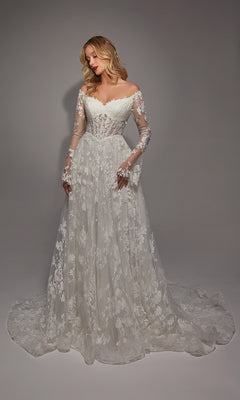 A woman models the Alyce 7150, a long ivory/sand dress with off-the-shoulder long sleeves, white lace, ruffle details on the fitted bodice, and a flowing skirt against a plain background.