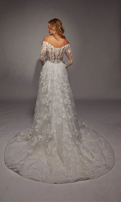 A woman faces away, wearing the Alyce 7150: a long white off-the-shoulder lace wedding dress with long sleeves, a flowing train, and delicate ruffle details.