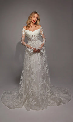 A woman poses in a studio wearing the Alyce 7150, a long white dress featuring off-the-shoulder long sleeves, white lace with ruffle details on the fitted bodice, and a flowing train.