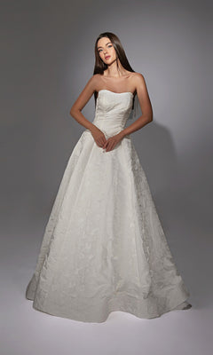 In a studio, a woman wears the Alyce 7155 long white dress in ivory, featuring a strapless design and delicate floral jacquard fabric that creates an elegant, floor-length silhouette.