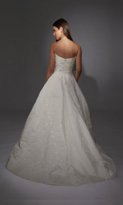 A woman wearing the Alyce 7155 long white strapless gown with elegant floral jacquard fabric stands facing away from the camera against a plain gray background.
