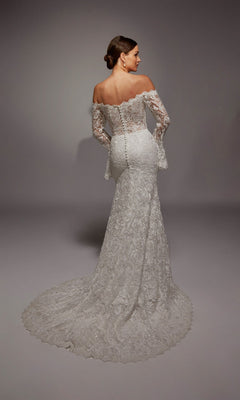 A woman stands against a plain background in the Alyce 7156: a long white, vintage-inspired bridal gown featuring an off-the-shoulder neckline, long lace sleeves, buttoned back, and a sweeping train.