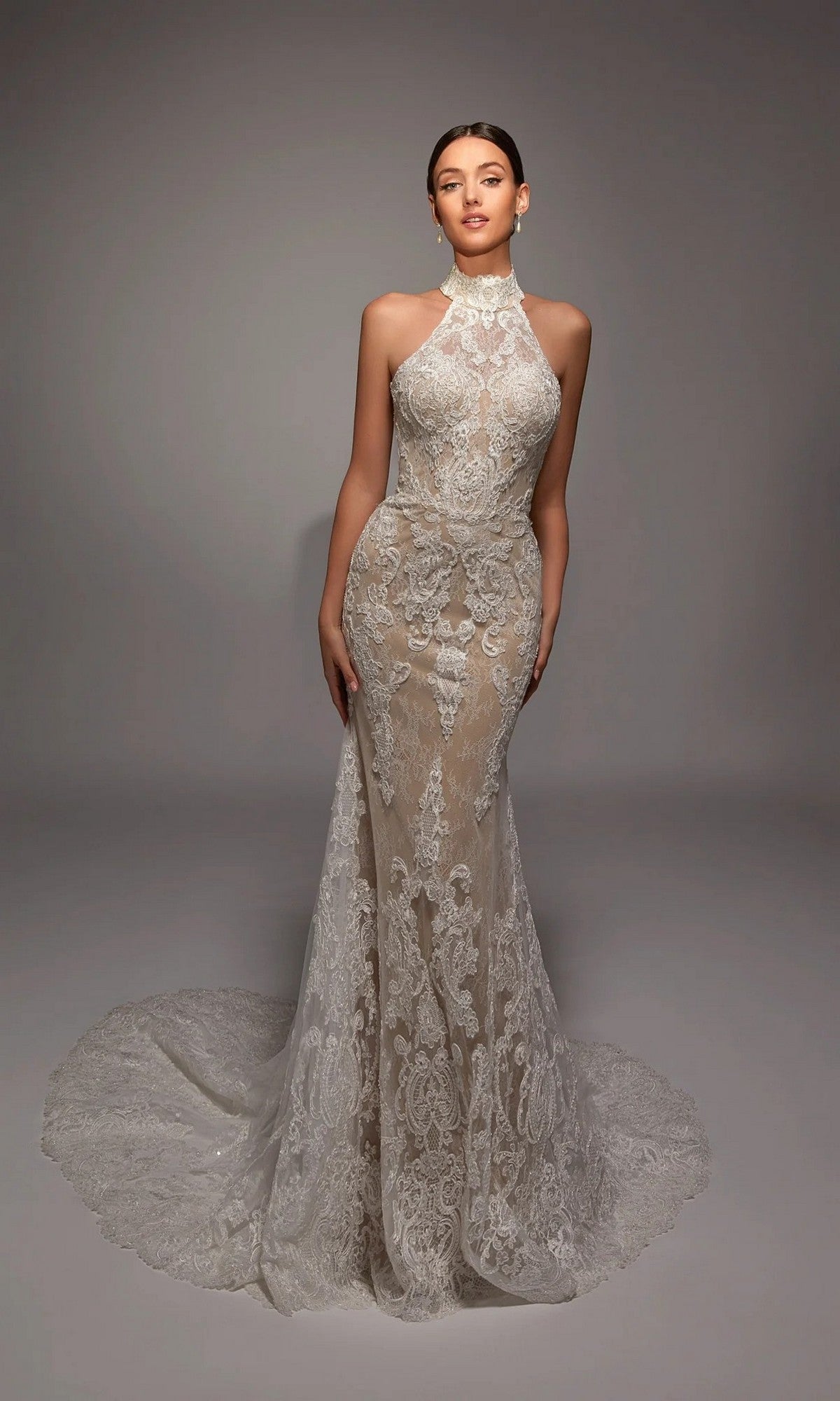 A woman stands against a plain studio backdrop wearing the Alyce 7159, a long white mermaid wedding gown featuring a sleeveless lace design, high neckline, and extended train.