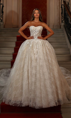 A woman stands on a red-carpeted staircase wearing the Alyce 7163 long white dress in Ivory/Sand, featuring a corset bodice and full skirt, with her hands on her hips.