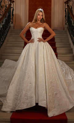 An elegant woman in the ivory Alyce 7164 long white dress stands on a red-carpeted staircase, wearing a tiara and posing with hands on hips.