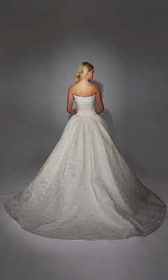 A woman stands facing away in the Alyce 7164 long white dress, a strapless gown with a full skirt and train, crafted from floral jacquard fabric, set against a neutral background.