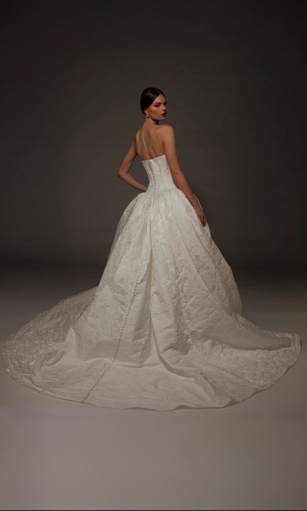 A woman stands in the Alyce 7165 long wedding dress, featuring a strapless sweetheart neckline and a full, ball gown skirt with a sweeping train, posing against a dark background.