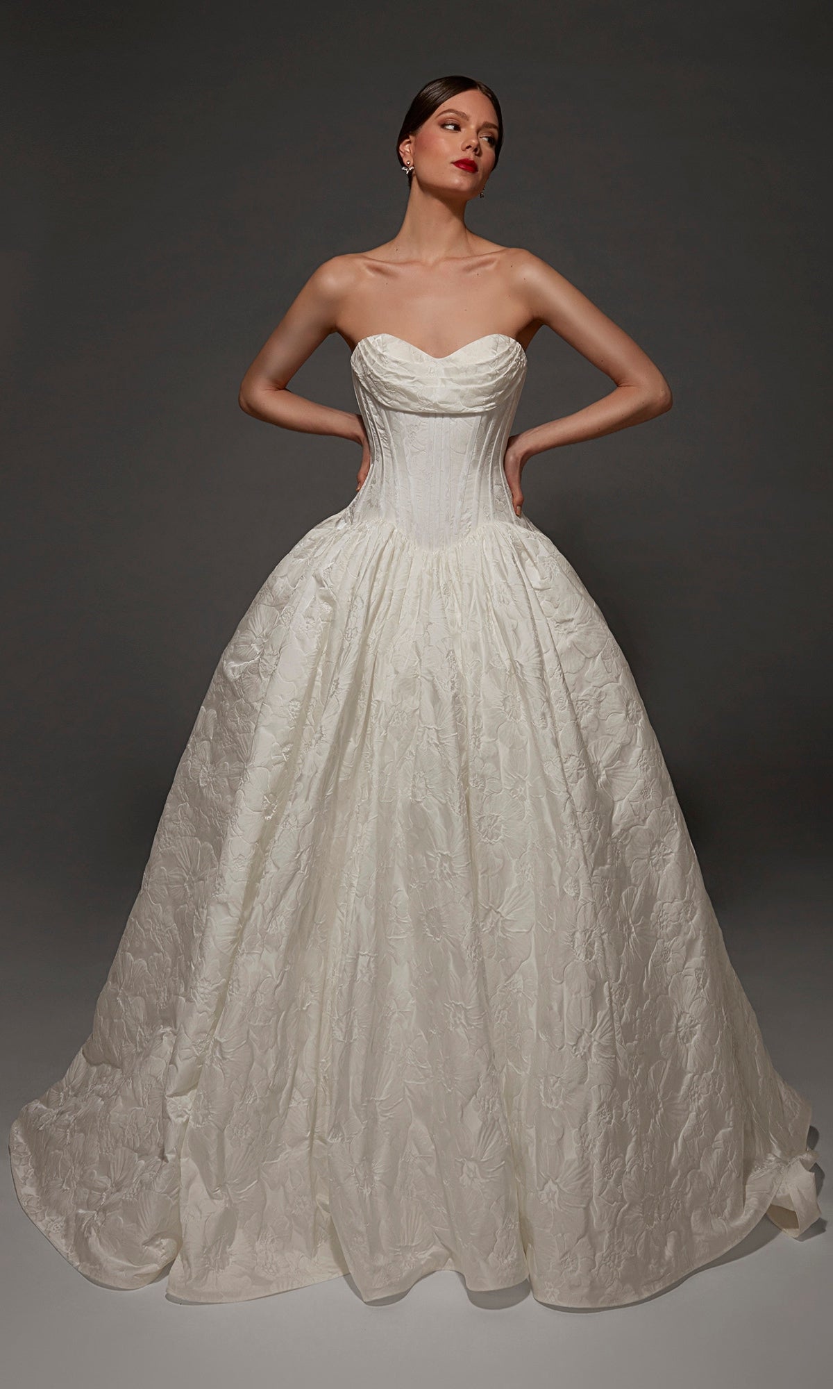 A woman wearing the Alyce 7165 long wedding dress, featuring a strapless sweetheart neckline, a fitted corset bodice, and a full textured jacquard ball gown skirt, stands against a dark background.