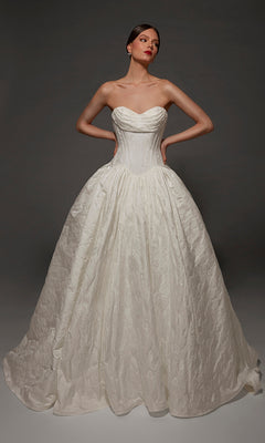 A woman wearing the Alyce 7165 long wedding dress, featuring a strapless sweetheart neckline, a fitted corset bodice, and a full textured jacquard ball gown skirt, stands against a dark background.