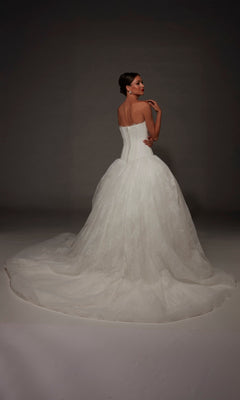 A woman wearing the Alyce 7169 Long Wedding Dress—a strapless white lace ball gown featuring a long train—stands facing away and looks over her shoulder against a gray backdrop.