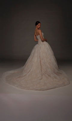 A woman wearing the Long Wedding Dress: Alyce 7169, featuring a strapless sweetheart neckline, lace detailing, a full ball gown skirt, and a long train, stands against a dark neutral background.