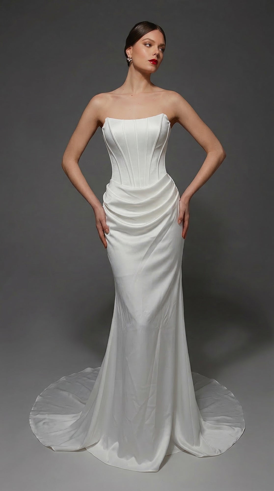 A woman wearing the Alyce 7170 long wedding dress in Diamond White, featuring a strapless satin design with a fitted corset bodice, draped waist detail, and an elegant long sheath skirt with a train, stands against a plain dark background.