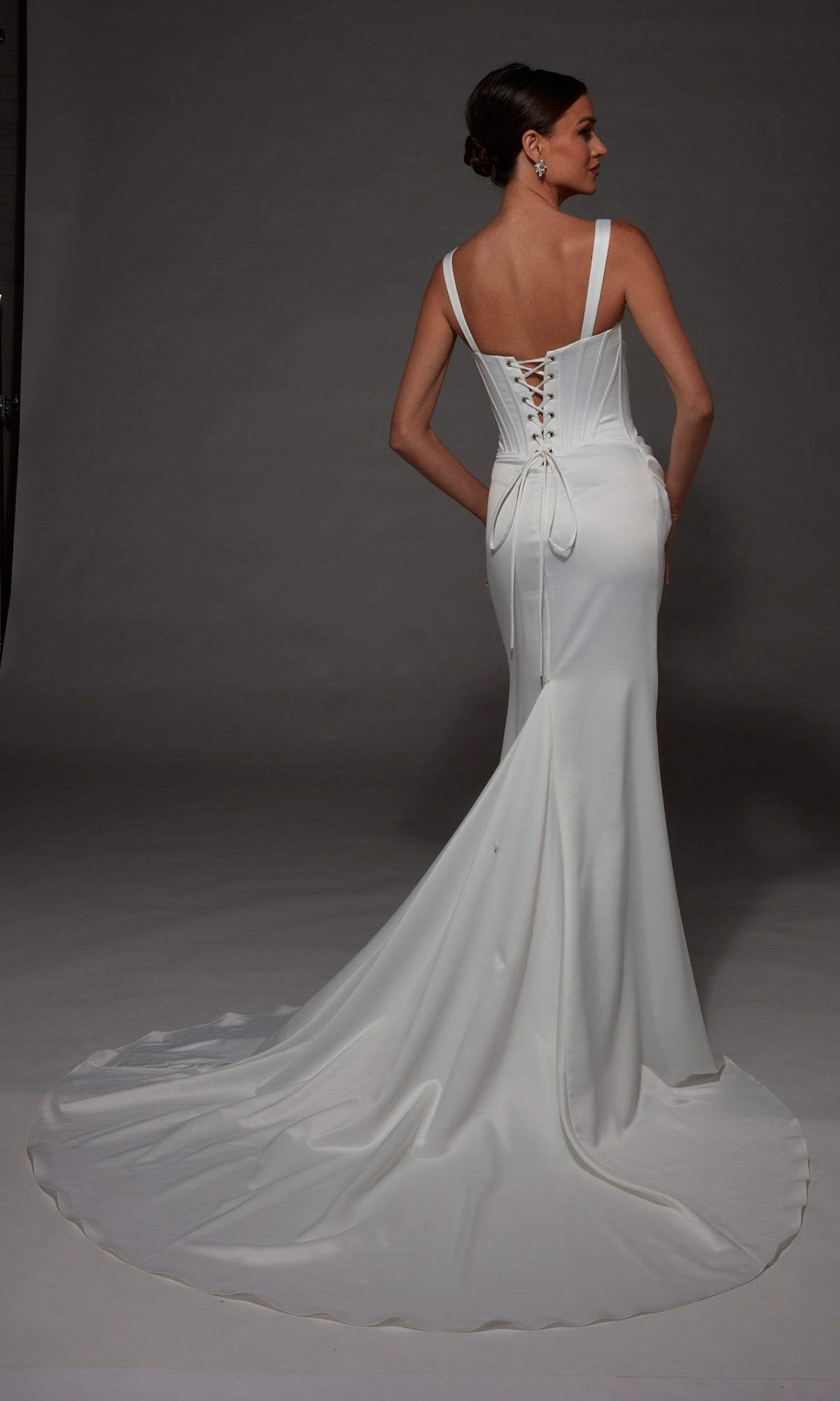 A woman stands facing away against a gray background, wearing the Alyce 7171 Long Wedding Dress—a fitted satin gown featuring a corset bodice, lace-up back, and a long train.