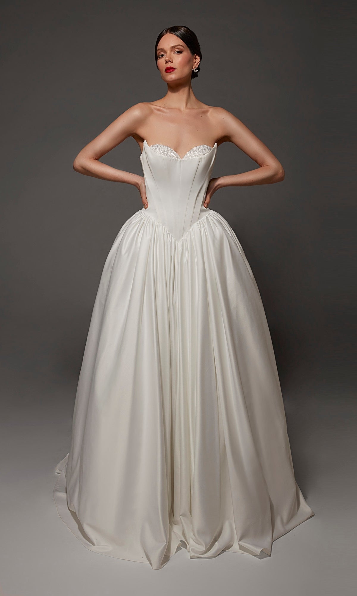 A woman poses in the Alyce 7172 long wedding dress, which features a strapless sweetheart neckline and fitted bodice; the satin ball gown’s full skirt flows elegantly against a plain dark background.