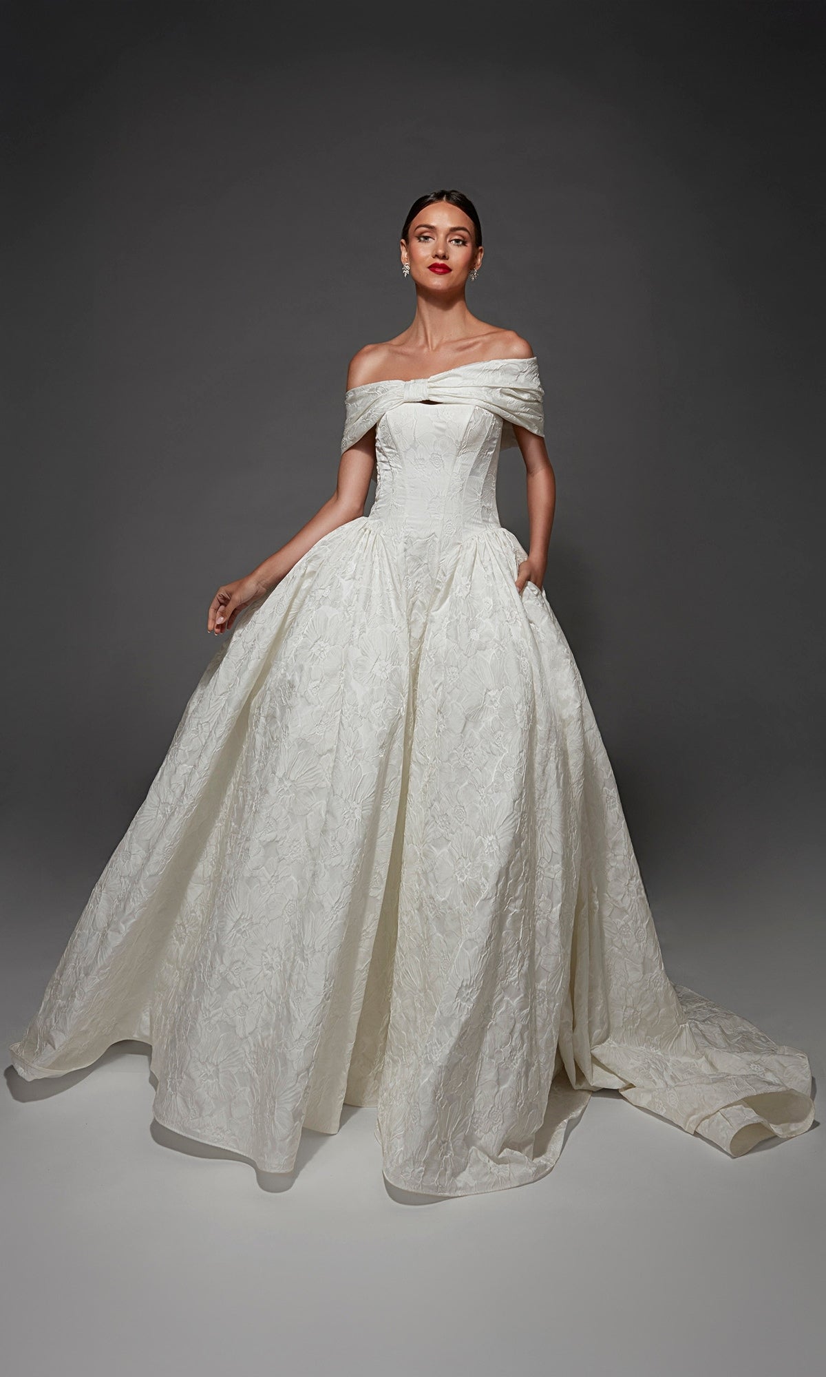 A woman stands against a plain dark background, wearing the Long Wedding Dress: Alyce 7174 in Diamond White—an off-the-shoulder jacquard ballgown featuring a strapless corset bodice and a textured, full skirt.