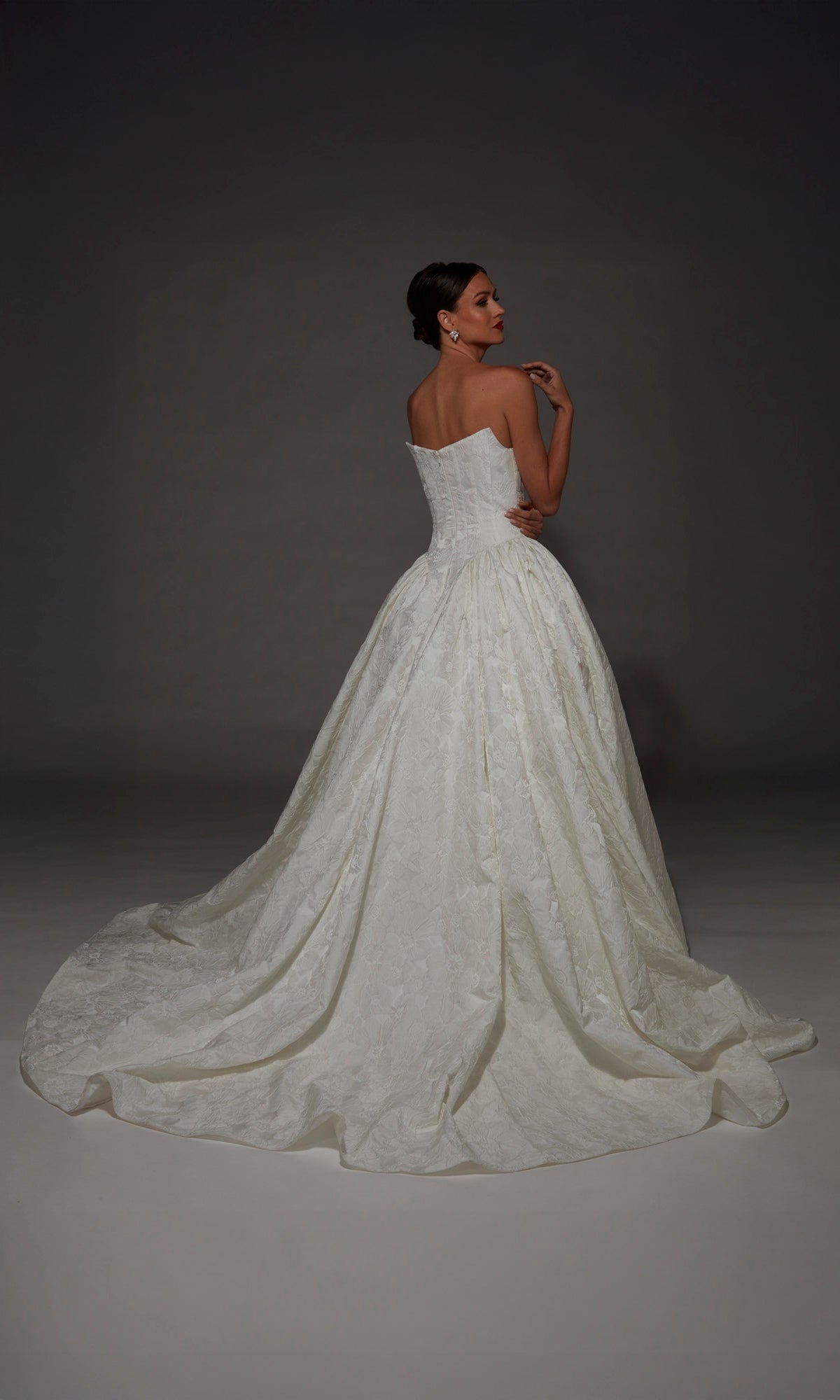 A woman stands against a dark background, facing away from the camera, wearing the Long Wedding Dress: Alyce 7174—a strapless white jacquard ballgown with a corset bodice, full skirt, and a long train.