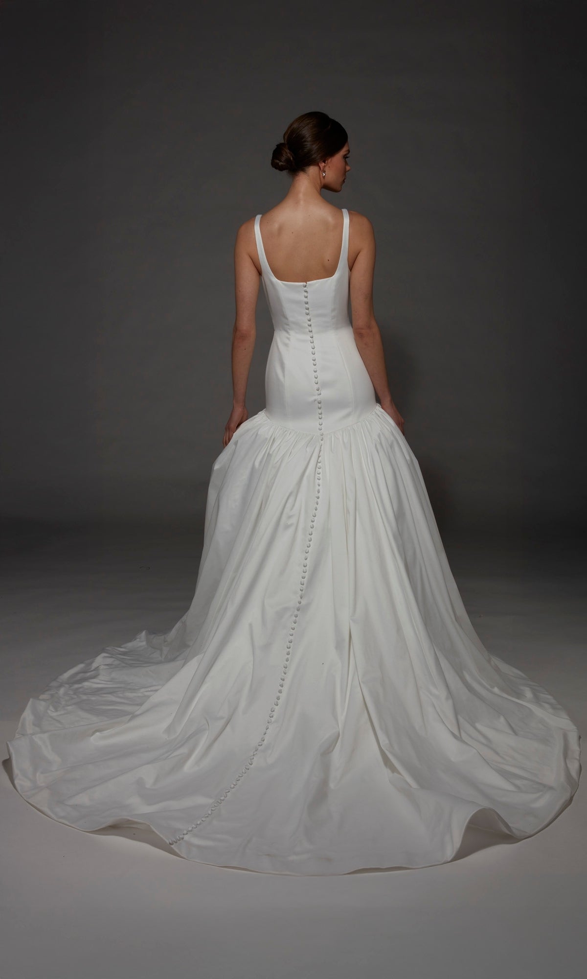 A woman stands facing away against a dark background, wearing the Alyce 7176 long wedding dress featuring a fitted bodice, sleeveless design, and elegant mermaid silhouette with a long train.