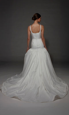A woman stands facing away against a dark background, wearing the Alyce 7176 long wedding dress featuring a fitted bodice, sleeveless design, and elegant mermaid silhouette with a long train.