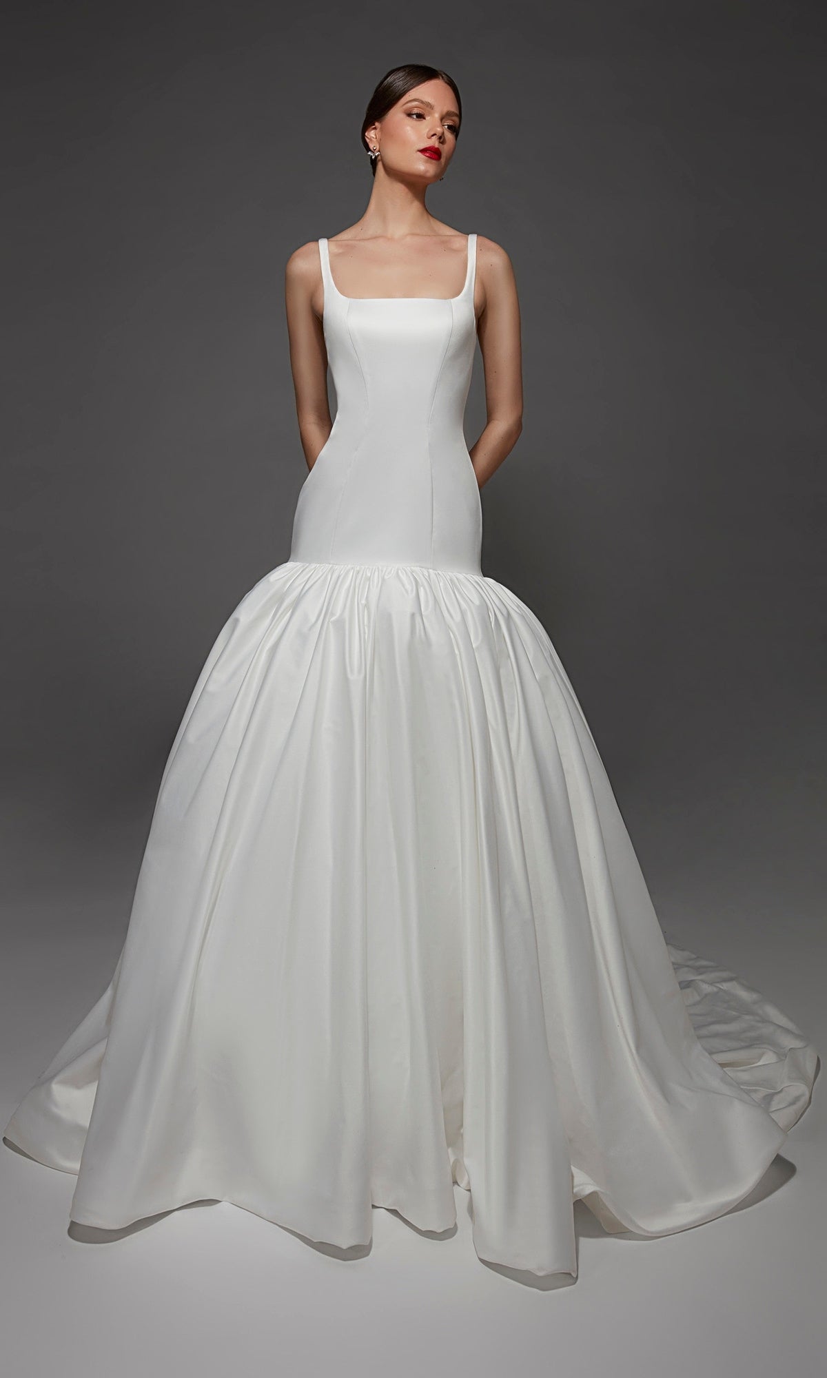 A woman wears the Alyce 7176 long wedding dress—a white mermaid gown featuring a fitted bodice and a voluminous skirt—as she stands against a gray background with her hands behind her back.
