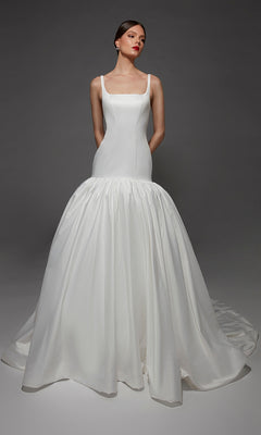 A woman wears the Alyce 7176 long wedding dress—a white mermaid gown featuring a fitted bodice and a voluminous skirt—as she stands against a gray background with her hands behind her back.