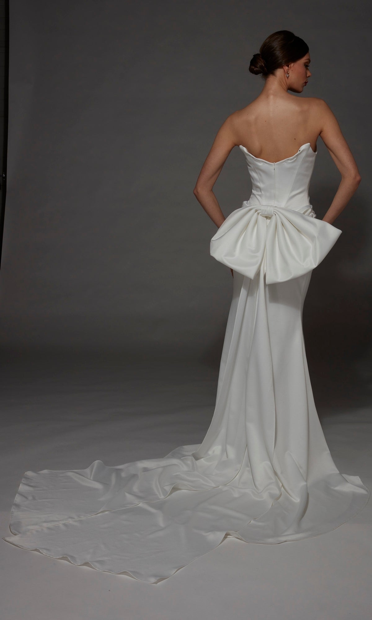 A woman wearing the Alyce 7177 long wedding dress, featuring elegant satin fabric, a strapless sweetheart neckline, and a large bow detail on the back, stands with her hands on her hips, facing away.