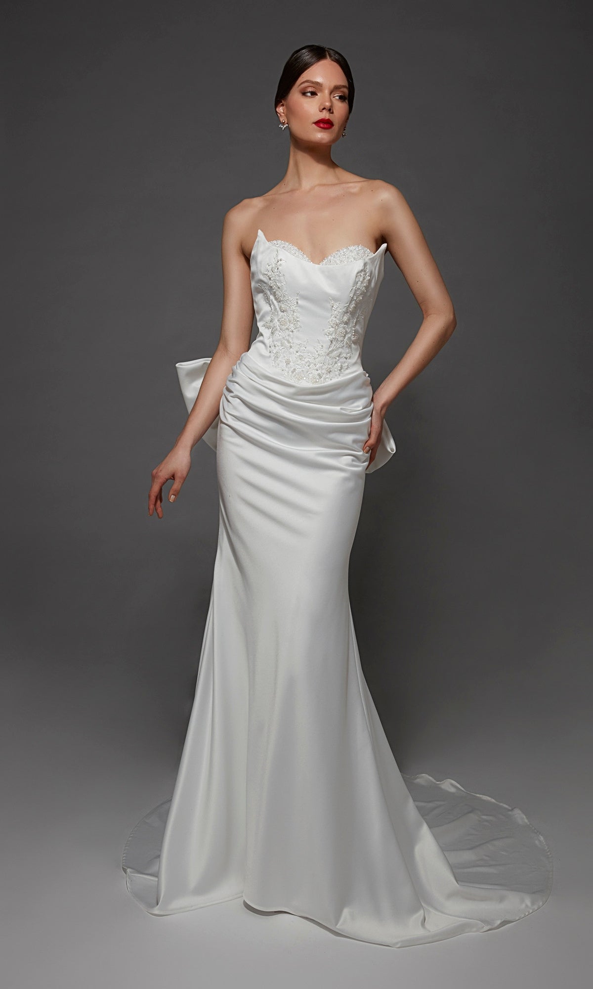 A woman wearing the Long Wedding Dress: Alyce 7177, a breathtaking bridal gown with a strapless sweetheart neckline, fitted bodice, delicate lace accents, and an elegant long train, stands against a dark gray background.