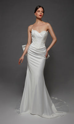 A woman wearing the Long Wedding Dress: Alyce 7177, a breathtaking bridal gown with a strapless sweetheart neckline, fitted bodice, delicate lace accents, and an elegant long train, stands against a dark gray background.