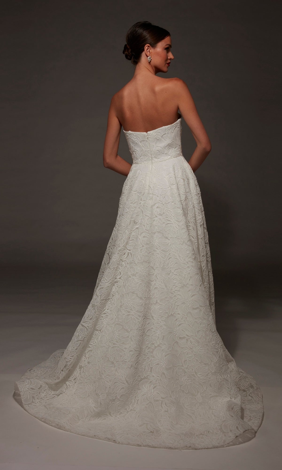 A woman faces away, showcasing the Long Wedding Dress: Alyce 7178—a strapless gown with a sweetheart neckline, floral textured pattern, fitted bodice, and a long, flowing chapel train—standing against a dark background.
