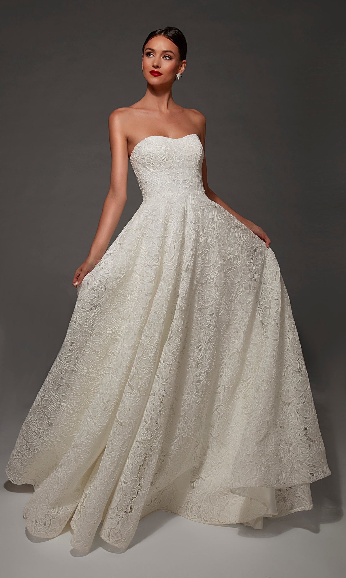 A woman poses against a dark background wearing the Alyce 7178 long wedding dress, featuring a strapless sweetheart neckline, a full skirt adorned with a textured floral pattern, and an elegant chapel train.