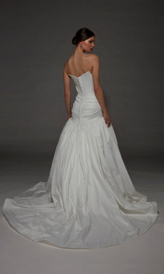 A woman stands with her back to the camera in the Alyce 7181 Long Wedding Dress, featuring a strapless satin design with a fitted bodice accented by buttons and a long, flowing train, set against a dark background.