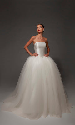 A woman wearing the Alyce 7182 Long Wedding Dress, featuring a strapless mikado bodice and a full, ball gown skirt, stands against a dark background with her hands on her hips.