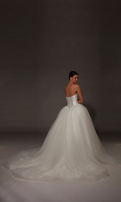 A woman stands facing away, looking over her shoulder, against a dark background, wearing the Alyce 7182 long wedding dress—a strapless white ball gown with a mikado bodice.