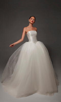A woman wearing the Long Wedding Dress: Alyce 7182 in Diamond White, featuring a strapless mikado bodice and a voluminous tulle skirt, stands against a dark background.