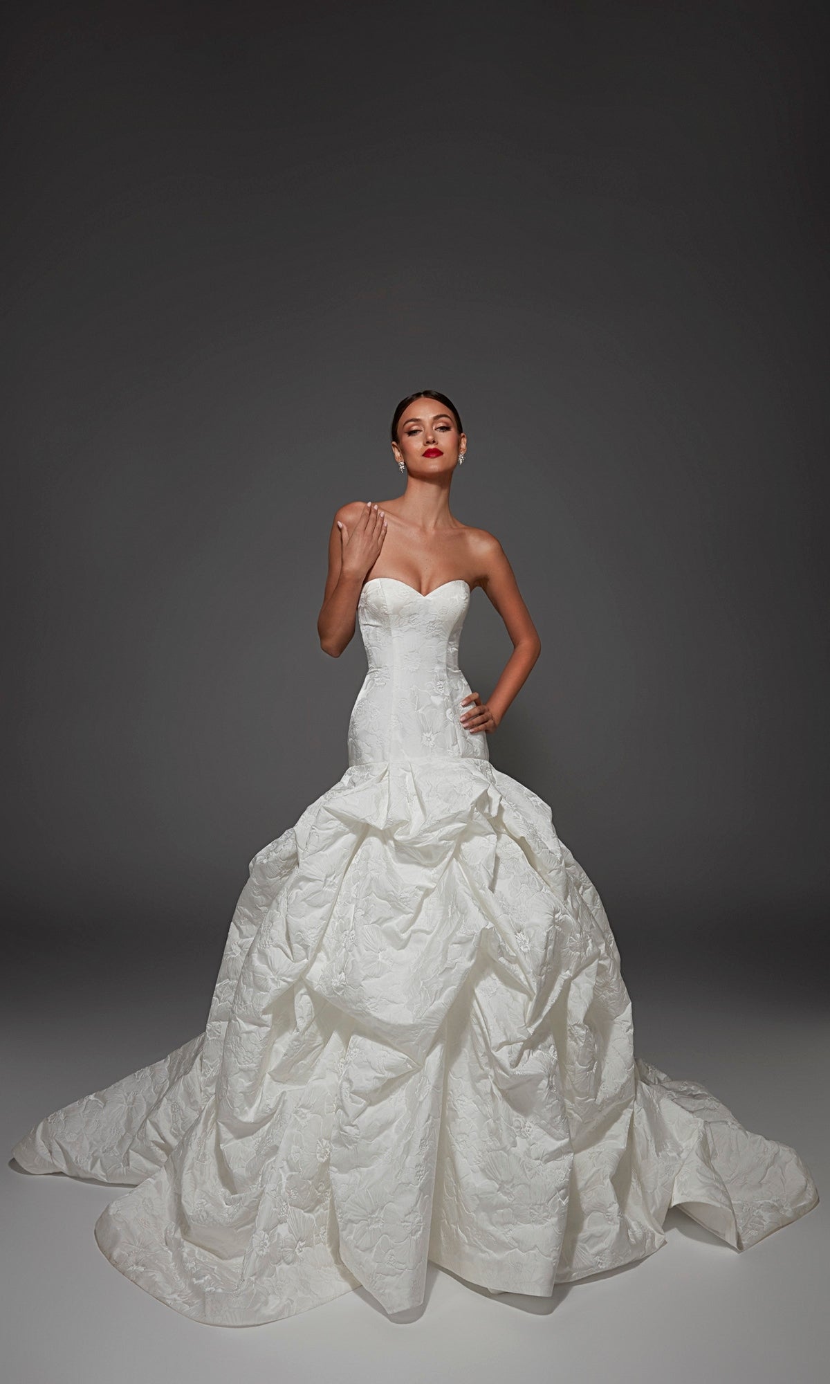 A woman poses against a dark, plain background wearing the Alyce 7185 long wedding dress in diamond white, featuring a strapless sweetheart neckline and a dramatic ruched ball-gown skirt.